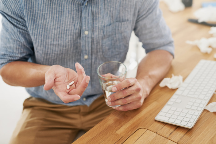 Mann sitzt an seinem Arbeitsplatz und nimmt eine Schmerztablette mit einem Glas Wasser ein