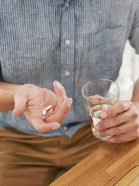 Mann sitzt an seinem Arbeitsplatz und nimmt eine Schmerztablette mit einem Glas Wasser ein