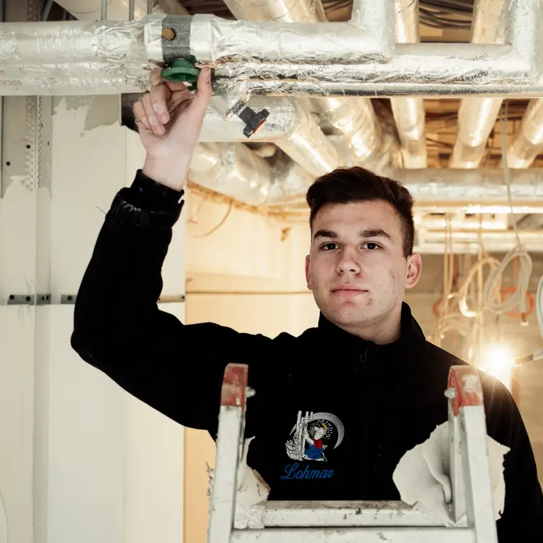 Junger Mann in Arbeitskleidung steht auf einer Leiter und prüft Rohre an der Decke