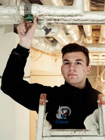 Junger Mann in Arbeitskleidung steht auf einer Leiter und prüft Rohre an der Decke