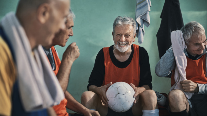 Ältere Männer sitzen lachend in Umkleidekabine, ein Mann hält einen Fußball in den Händen