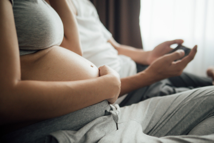 Schwangere und ihr Mann sitzen auf einem Sofa, er checkt sein Smartphone