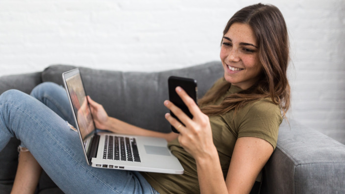 Junge Frau sitzt auf dem Sofa mit Handy und Laptop.