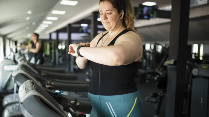 Eine Frau steht auf einem Laufband im Fitness-Studio und schaut auf ihre Smartwatch. 