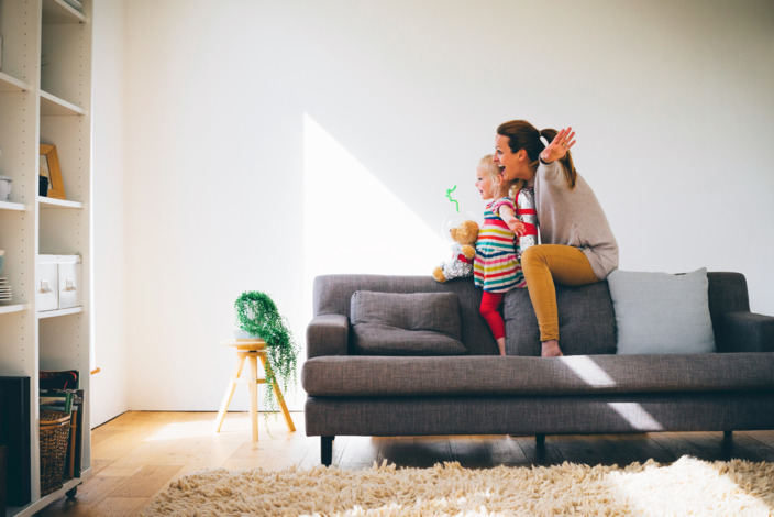 Mutter und Tochter sitzen auf der Rückenlehne eines grauen Sofas und spielen Flugzeug