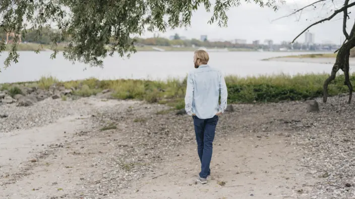 Junger Mann geht gedankenversunken am Ufer eines Flusses entlang.