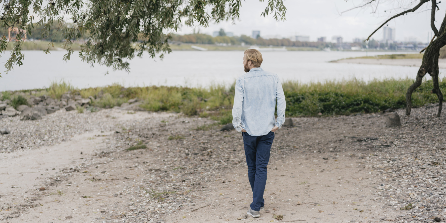 junger Mann geht gedankenversunken am Ufer eines Flusses entlang