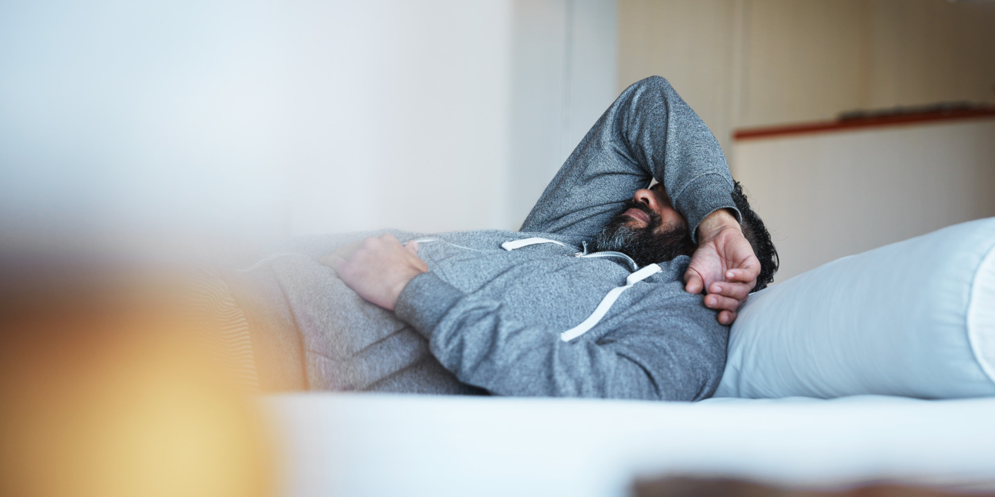 Mann in grauem Pullover liegt in Bett und legt seinen Arm über sein Gesicht. 