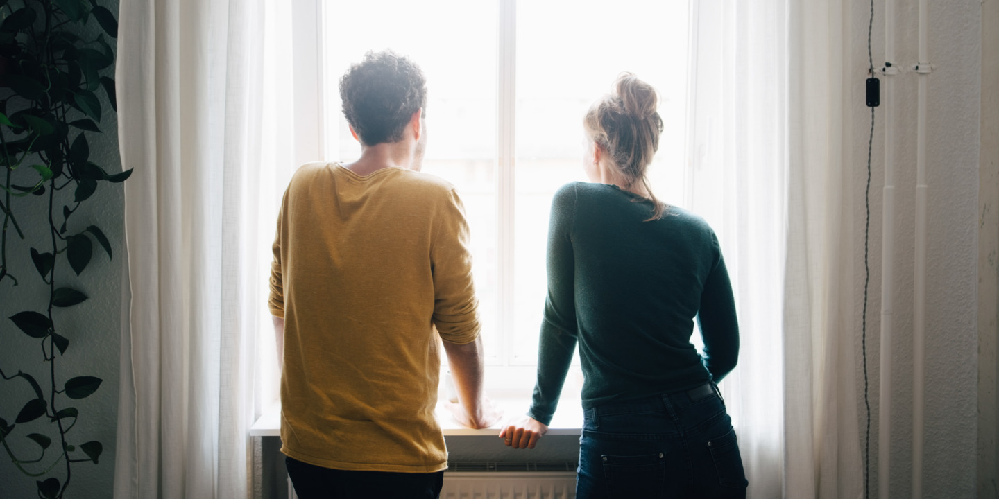 Junges Paar steht mit dem Rücken zum Betrachter und schaut aus einem Fenster.