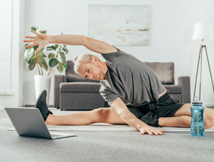 Mann sitzt im Wohnzimmer und macht eine Dehnübung vor dem aufgeklappten Laptop