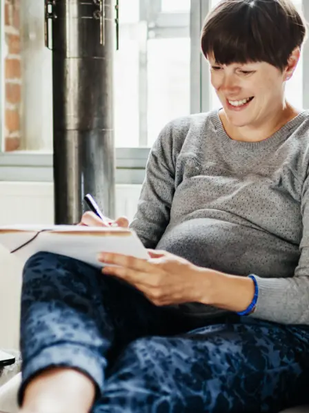 Schwangere Frau sitzt mit Stift und Block auf dem Sofa, während ein Kind neben ihr spielt