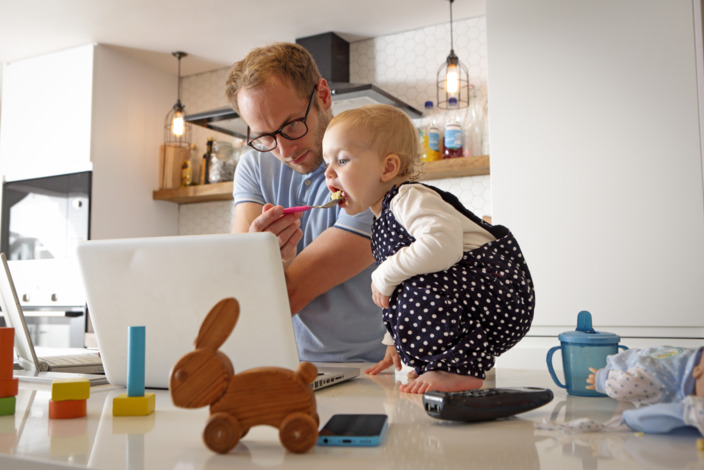 Vater füttert seinen kleinen Sohn und arbeitet gleichzeitig an seinem Laptop