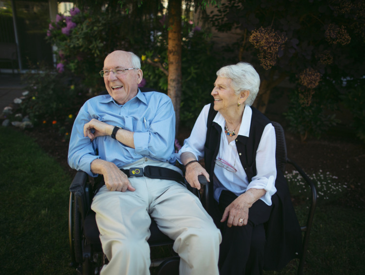 Seniorin und ihr pflegebedürftiger Mann sitzen gut gelaunt im Garten
