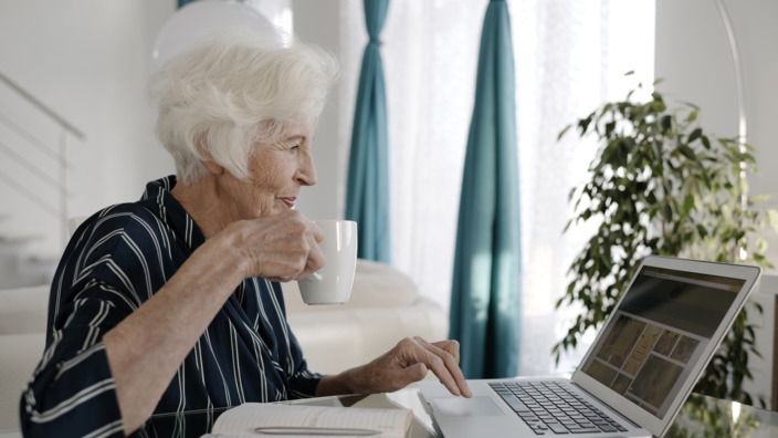 Seniorin sitzt mit einer Tasse Tee vor dem Laptop und prüft ihre Unterlagen