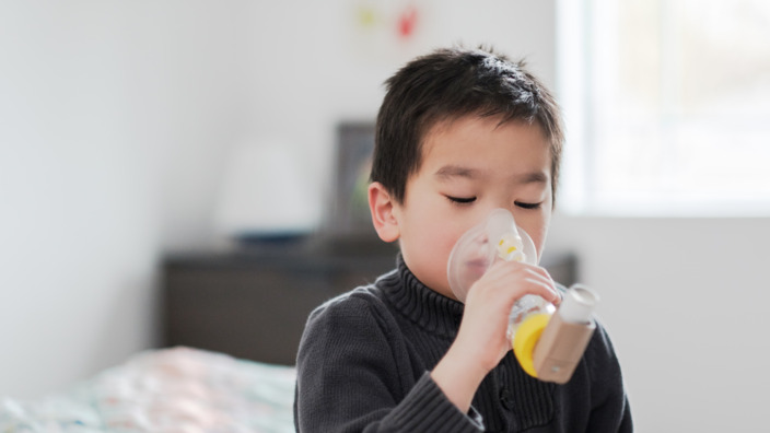 Junge sitzt auf dem Bett und inhaliert mithilfe eines Inhalators