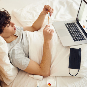 Ein junger Mann misst Fieber mit Thermometer im Bett und spricht per Laptop in einer Telemedizin-Video-Sprechstunde mit einem Arzt