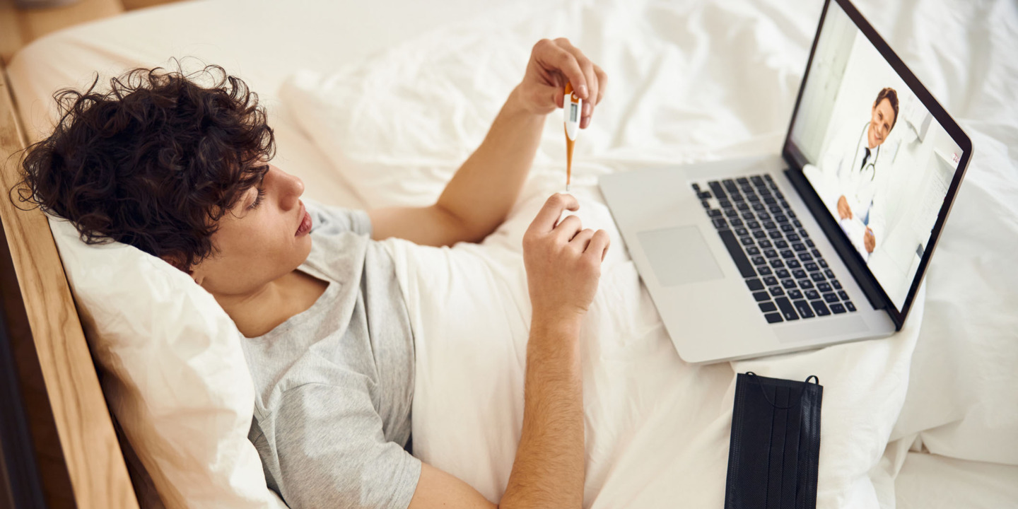 junger Mann liegt im Bett und hat eine Videosprechstunde beim Arzt