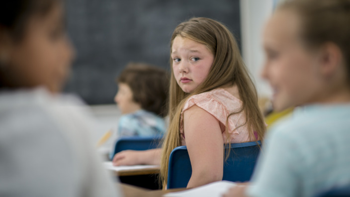 Mädchen sitzt in einem Klassenraum und schaut ängstlich hinter sich