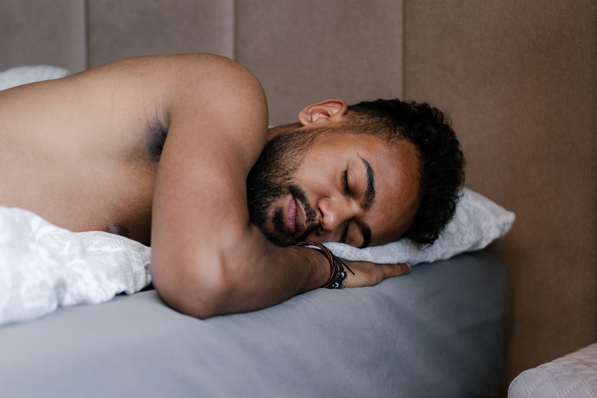 Ein Mann schläft seitlich auf Kissen in einem Bett, entspannter Gesichtsausdruck und nackter Oberkörper sichtbar.