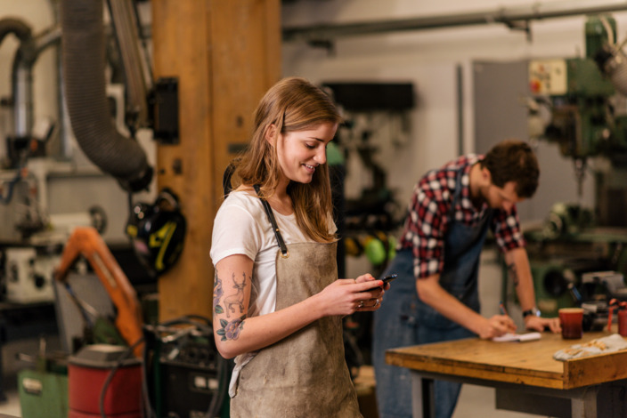 Junge Auszubildene steht in der Werkstatt und schaut auf ihr Handy