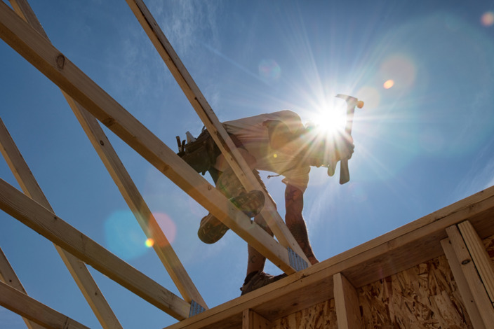 Ein Handwerker arbeitet an dem Dach eines Holzhauses mit einem Hammer in der starken Mittagssonne