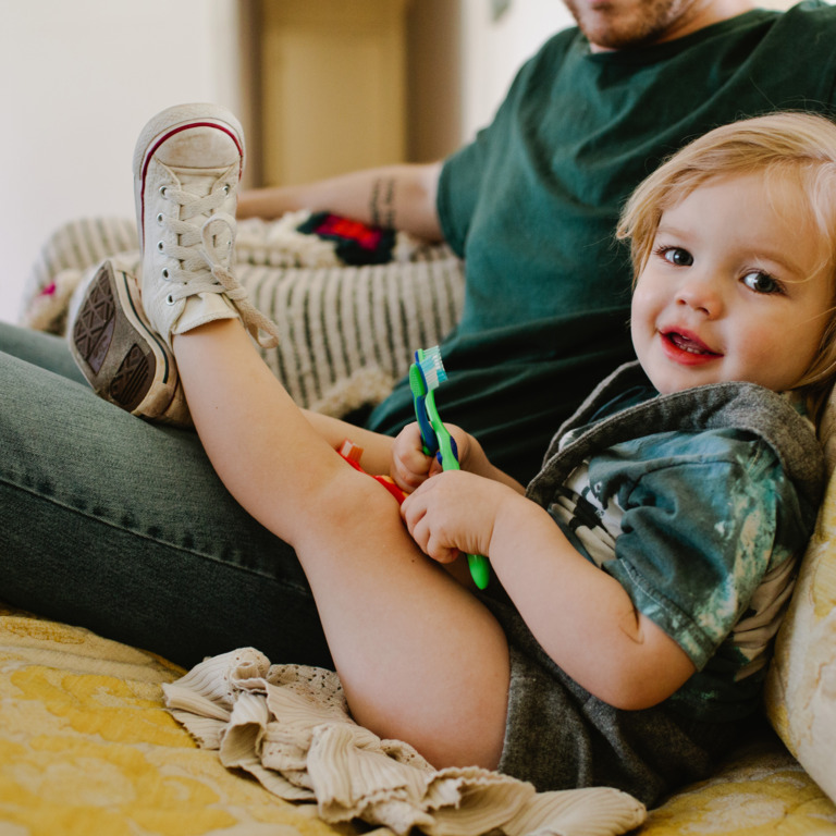 kleiner Junge sitzt auf der Couch und spielt mit verschiedenen Kinderzahnbürsten